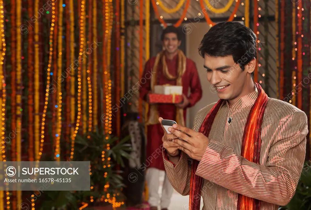 Man text messaging on a mobile phone with his friend holding gifts on Diwali