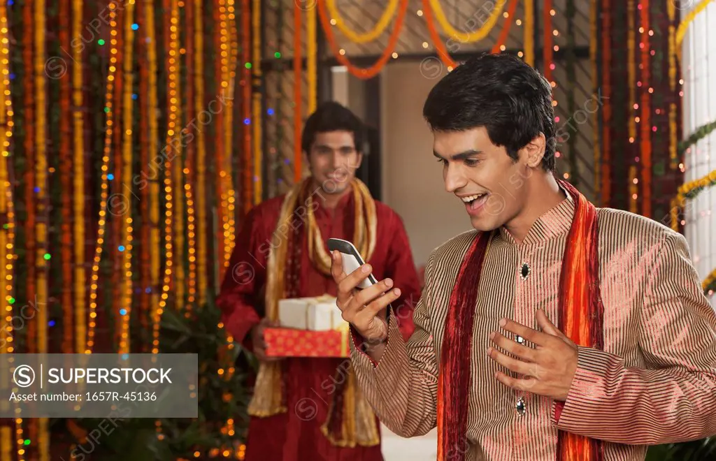 Man text messaging on a mobile phone with his friend holding gifts on Diwali