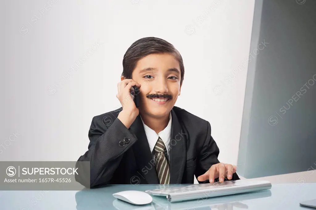 Boy imitating like businessman talking on a mobile phone while working on a desktop pc