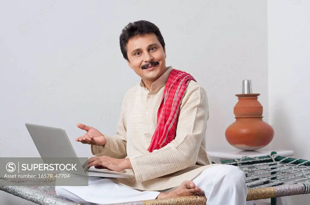 Portrait of a farmer sitting on a cot and using a laptop