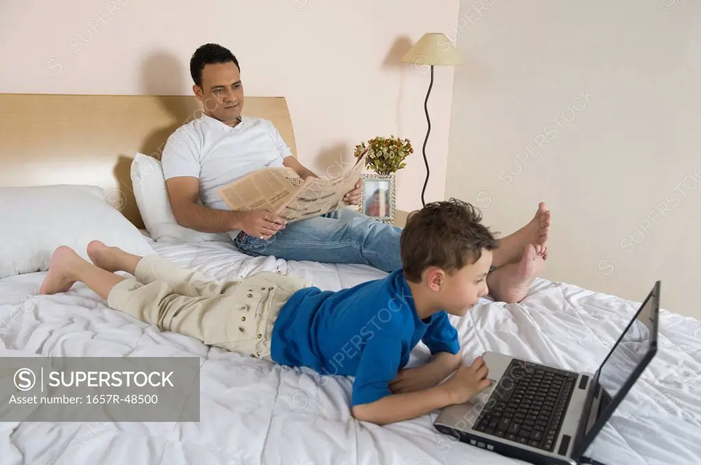 Boy using a laptop with his father reading a newspaper beside him