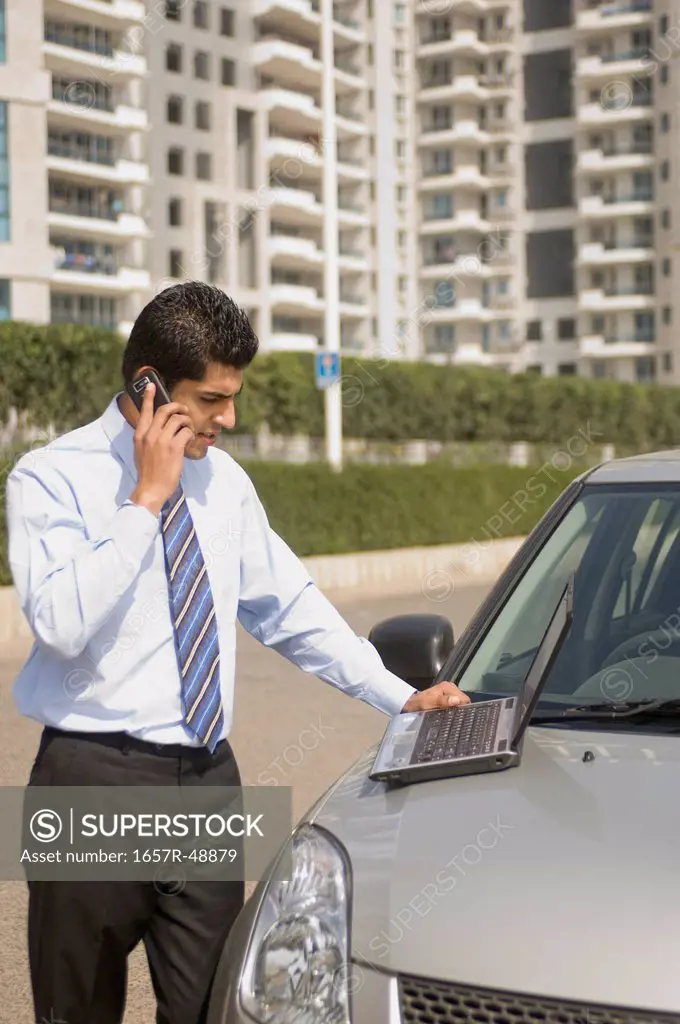 Businessman talking on a mobile phone while using a laptop on car bonnet, Gurgaon, Haryana, India