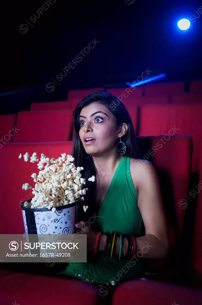 Woman holding popcorns while watching movie and looking surprised in a cinema hall