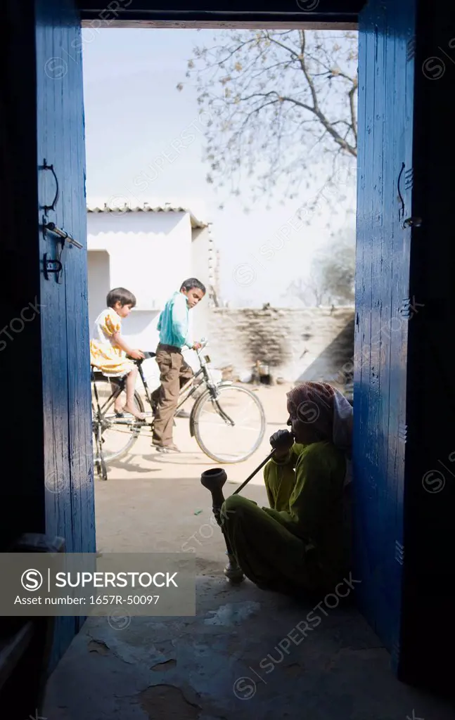 Woman smoking a hookah pipe with children riding a bicycle, Farrukh Nagar, Gurgaon, Haryana, India