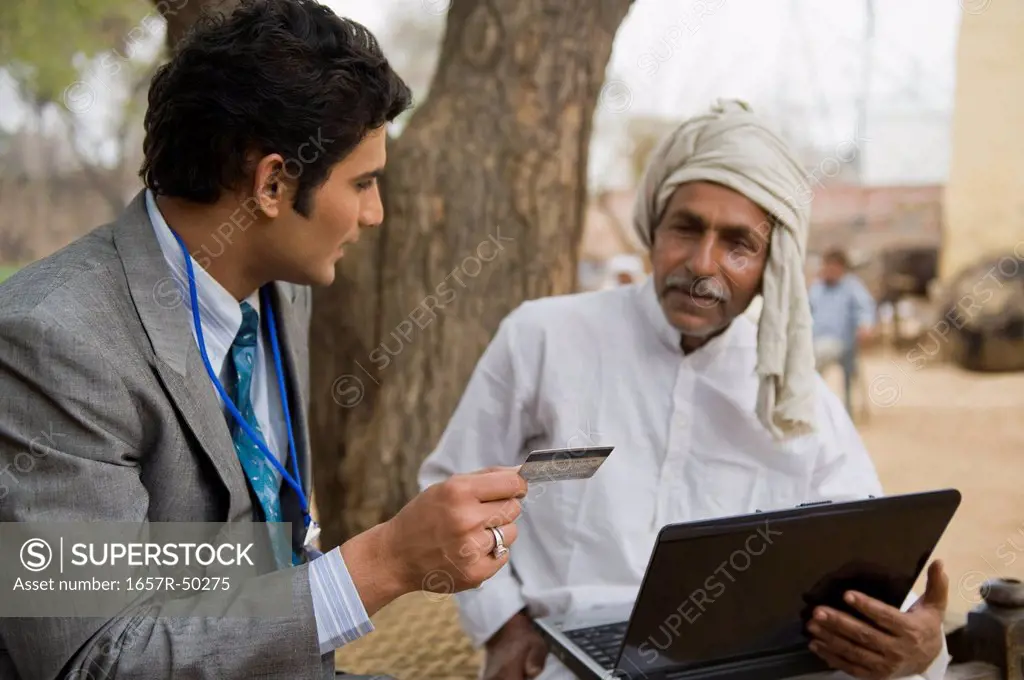 Businessman holding a credit card sitting with a farmer holding a laptop, Hasanpur, Haryana, India