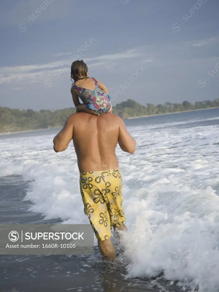 Rear view of a man carrying his daughter on his shoulders and walking on the beach