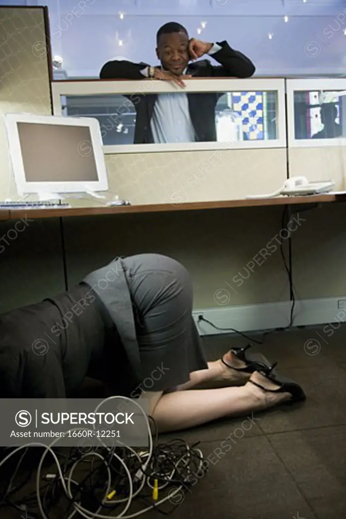 Businesswoman crouching under a computer desk with a businessman looking at her