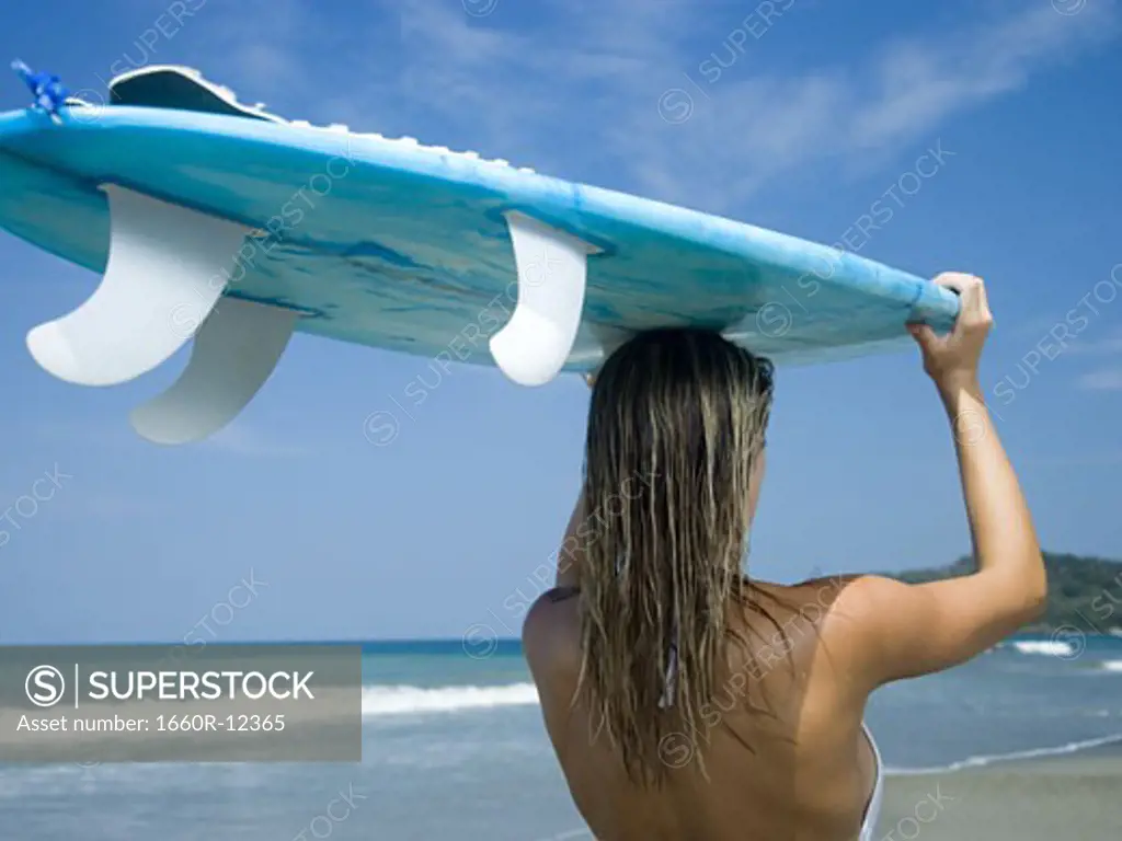 Rear view of a young woman holding a surfboard over her head