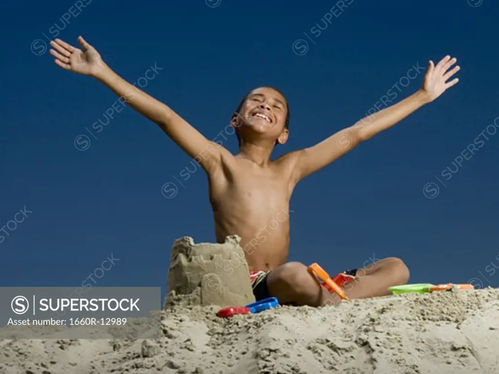 Low angle view of a boy sitting beside a sand castle with his arms outstretched