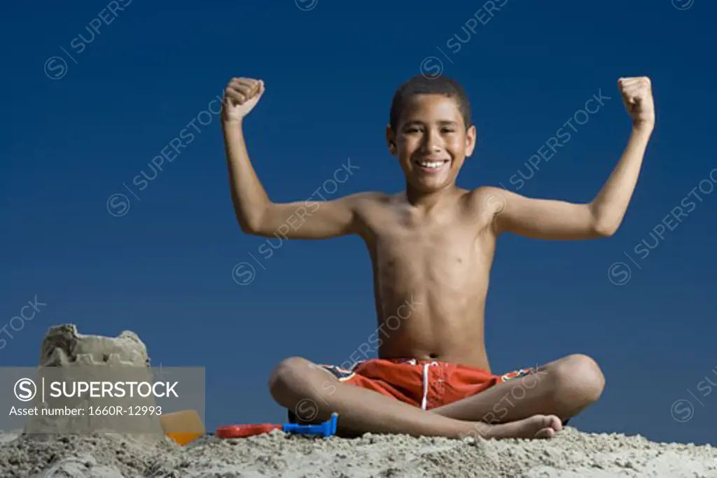 Portrait of a boy sitting near a sand castle and flexing his muscles