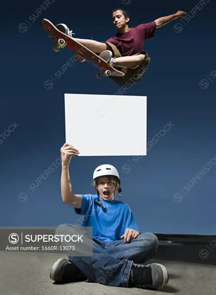 Low angle view of a teenage boy jumping with a skateboard over another teenage boy holding a blank sign