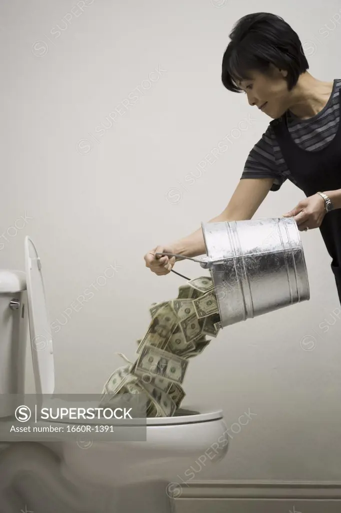Mid adult woman emptying a bucket of currency notes into a toilet bowl