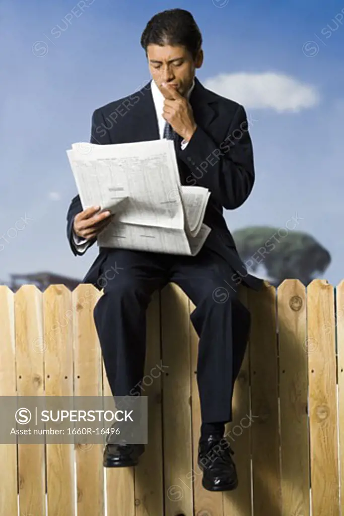 Businessman sitting on fence reading newspaper
