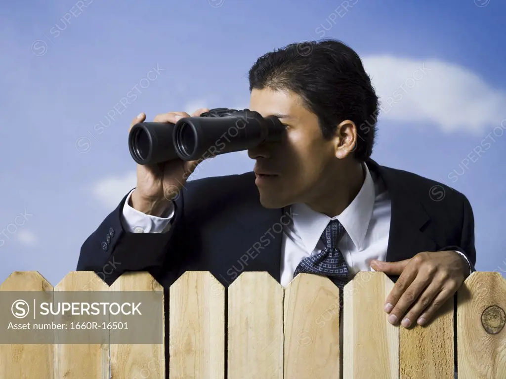 Businessman looking through binoculars over wooden fence