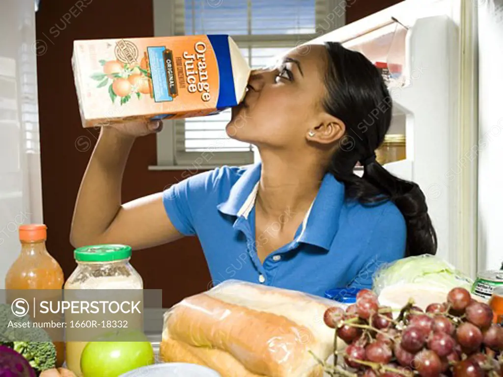 Woman drinking orange juice from carton in refrigerator with healthy foods