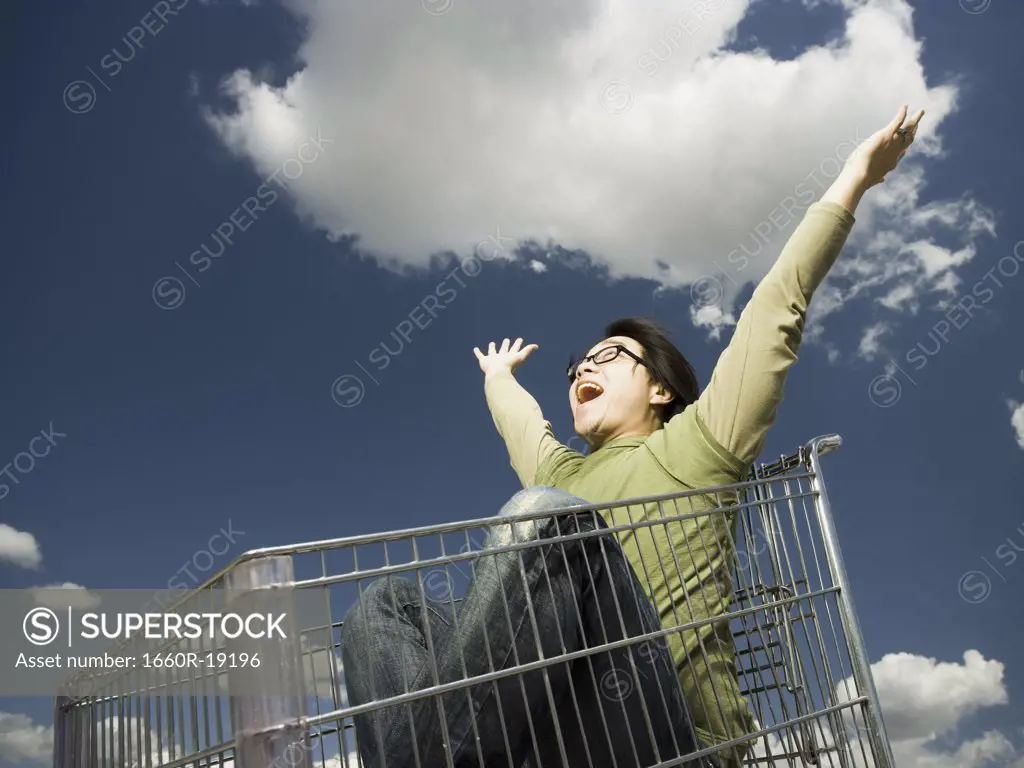 Man with eyeglasses in shopping cart outdoors with arms up smiling