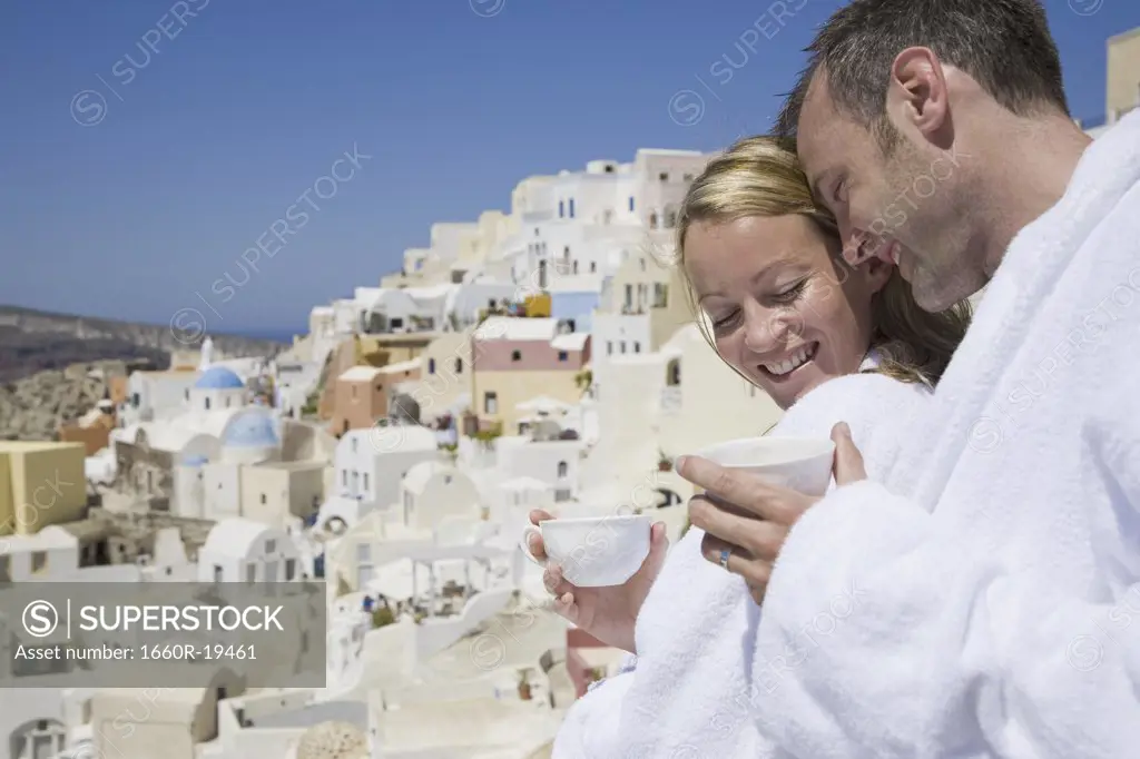 Couple in bathrobes with cups smiling with scenic village in background