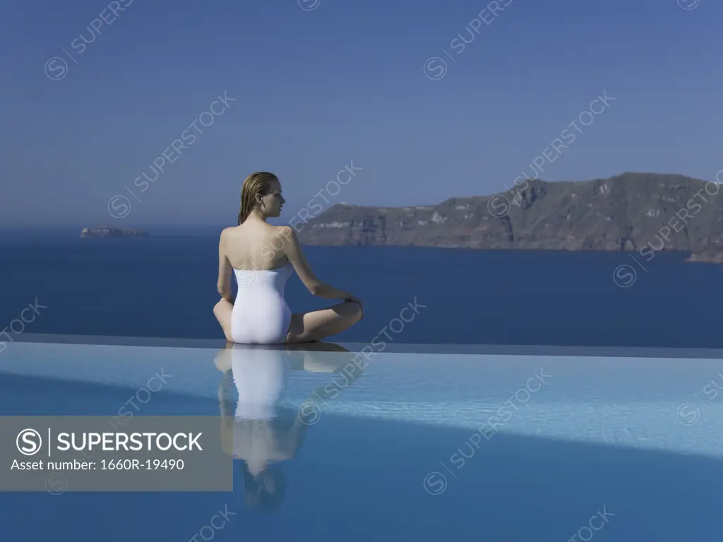 Woman in swimsuit sitting cross legged by infinity pool outdoors
