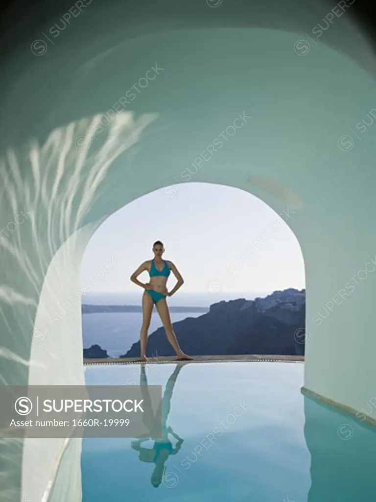 Woman in bikini standing at edge of infinity pool outdoors with arch and rock formation