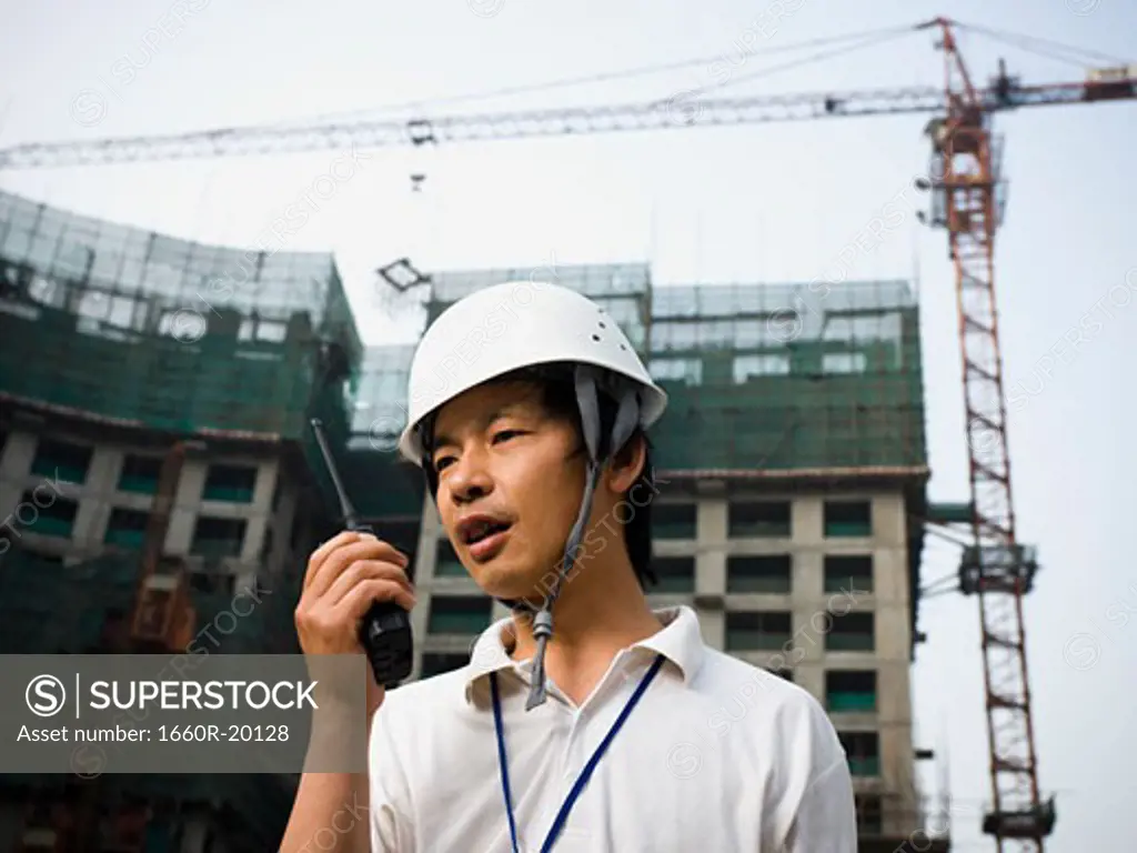Man with construction helmet and crane in background holding walkie talkie