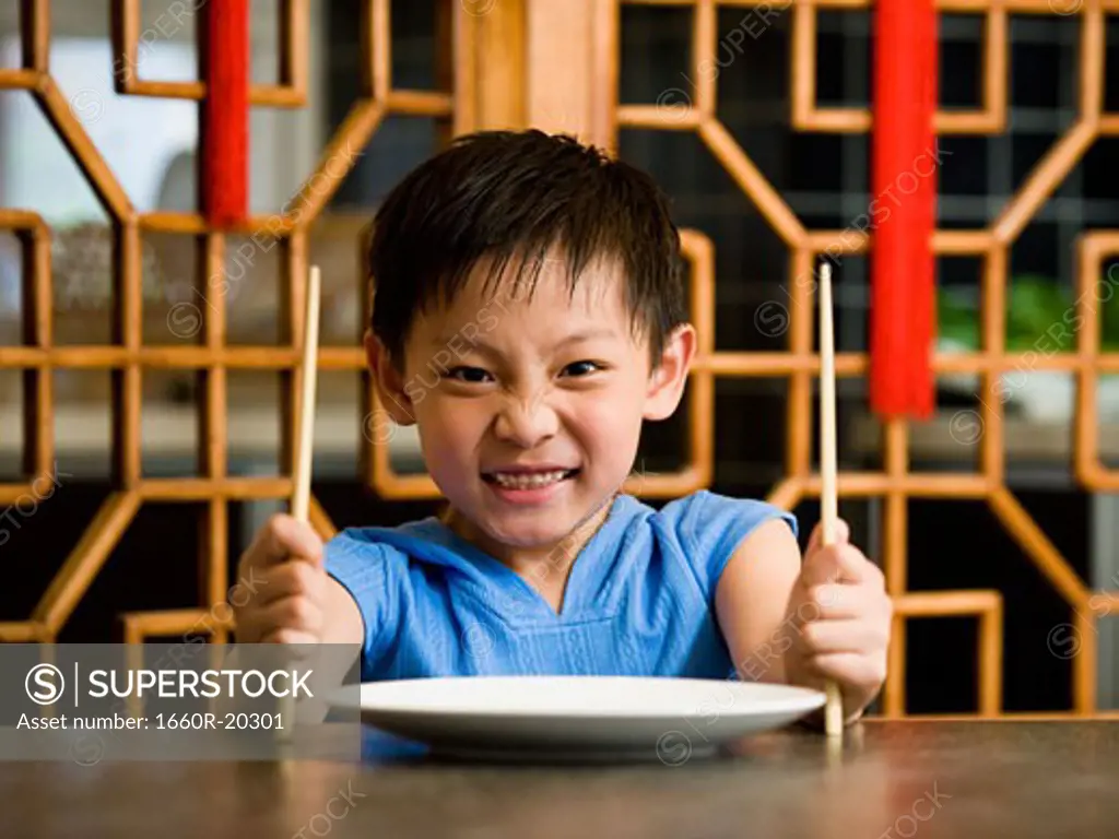Boy sitting at table with empty plate holding chopsticks scowling
