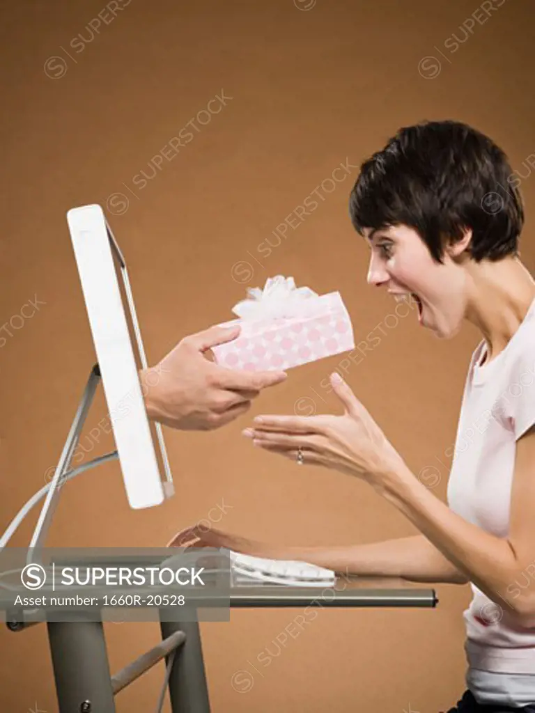 Woman sitting at computer with hand emerging from monitor holding gift box