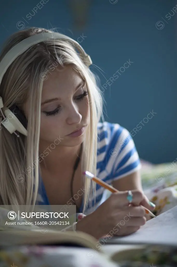 Close up of concentrating teenage girl with long blonde hair looking down and writing homework on bed and listening to headphones