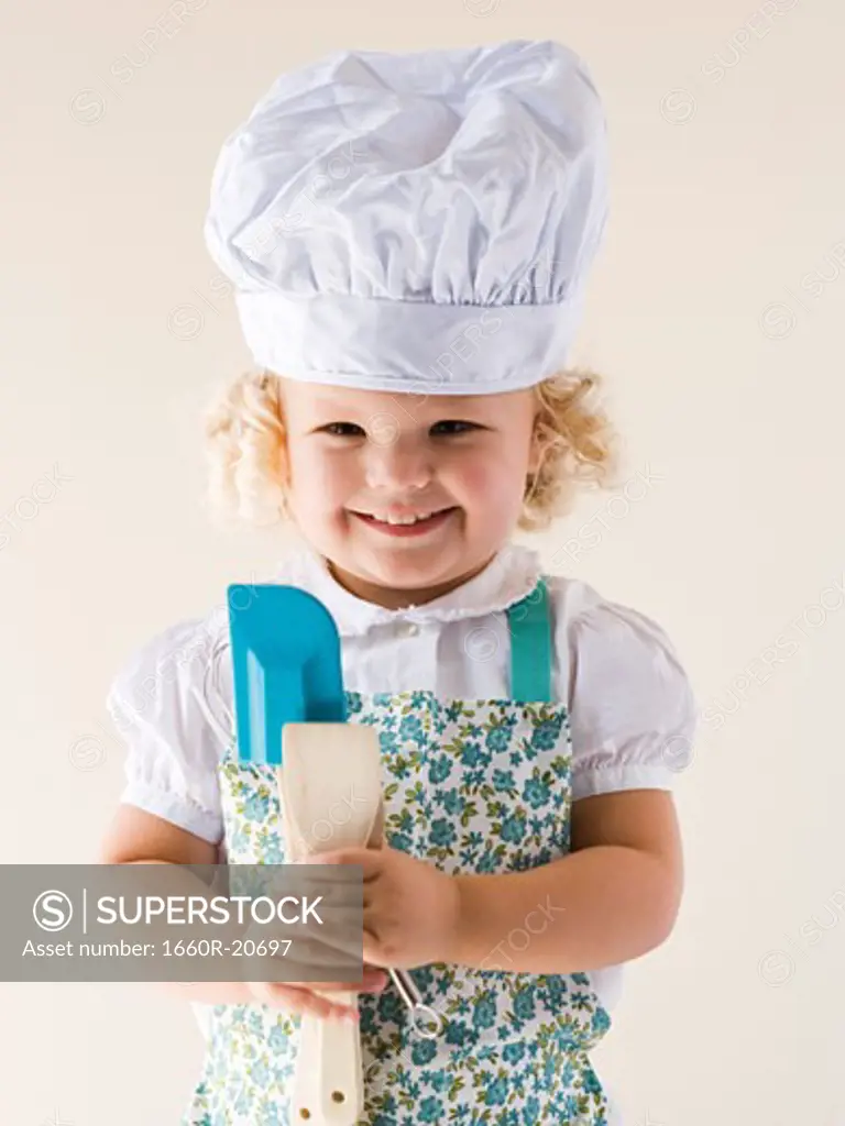 Little girl wearing a chef's hat and holding spoons.