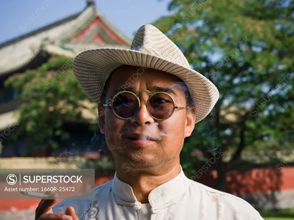 Man with sunglasses and straw hat smoking cigarette outdoors with pagoda in background