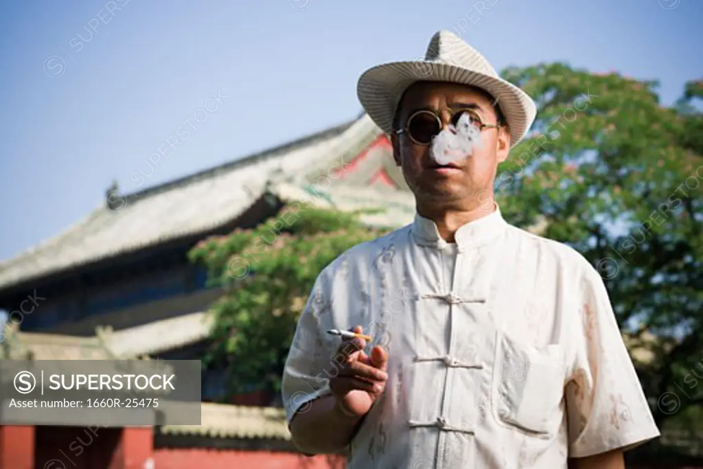 Man with sunglasses and straw hat smoking cigarette outdoors with pagoda in background