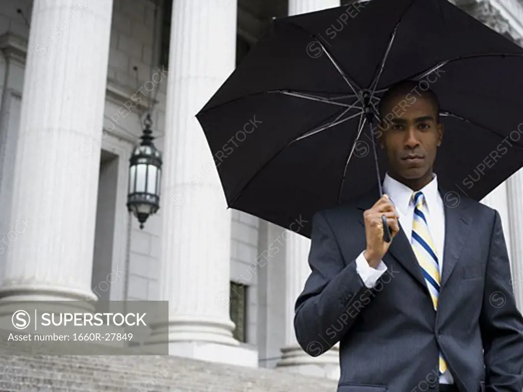 Portrait of a male lawyer standing in front of a courthouse and holding an umbrella