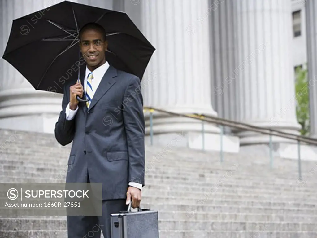 Portrait of a male lawyer standing in front of a courthouse and holding an umbrella