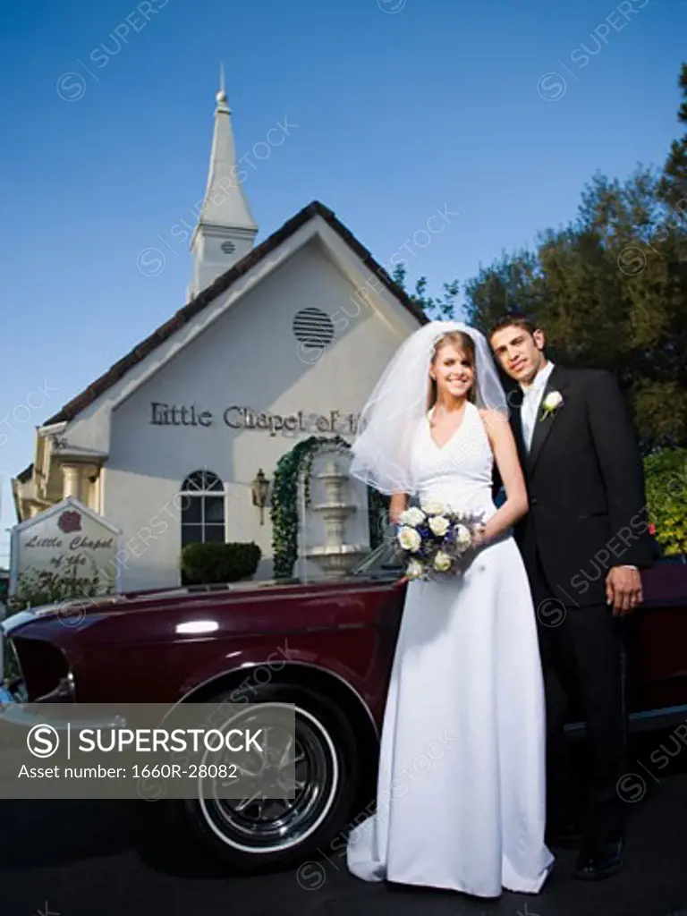 Portrait of a newlywed couple standing near a car in front of a chapel
