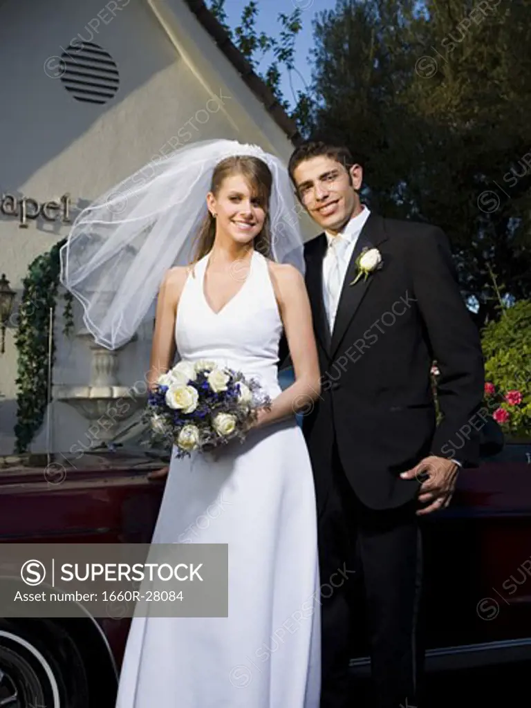 Portrait of a newlywed couple standing near a car in front of a chapel