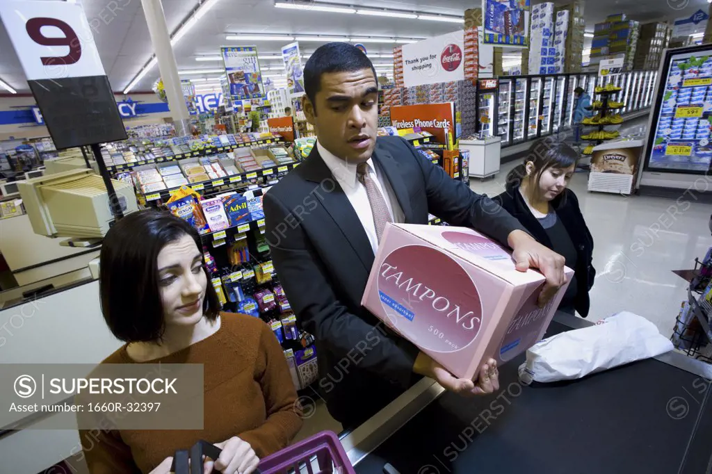 Man at grocery checkout with box of tampons and two women