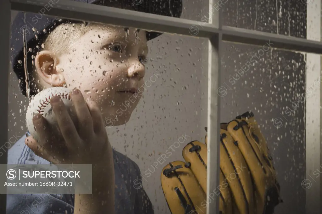 Young boy with baseball glove looking out window on rainy day