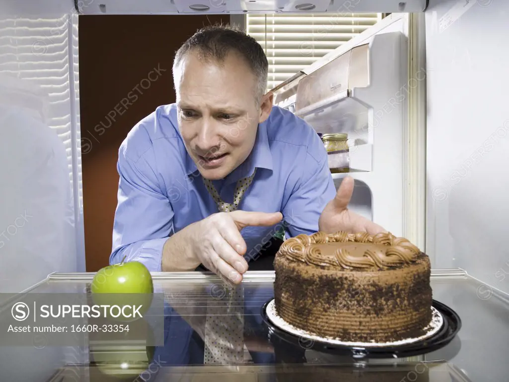 Man in refrigerator reaching for green apple looking at chocolate cake