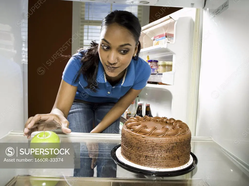 Woman in refrigerator with apple looking at chocolate cake