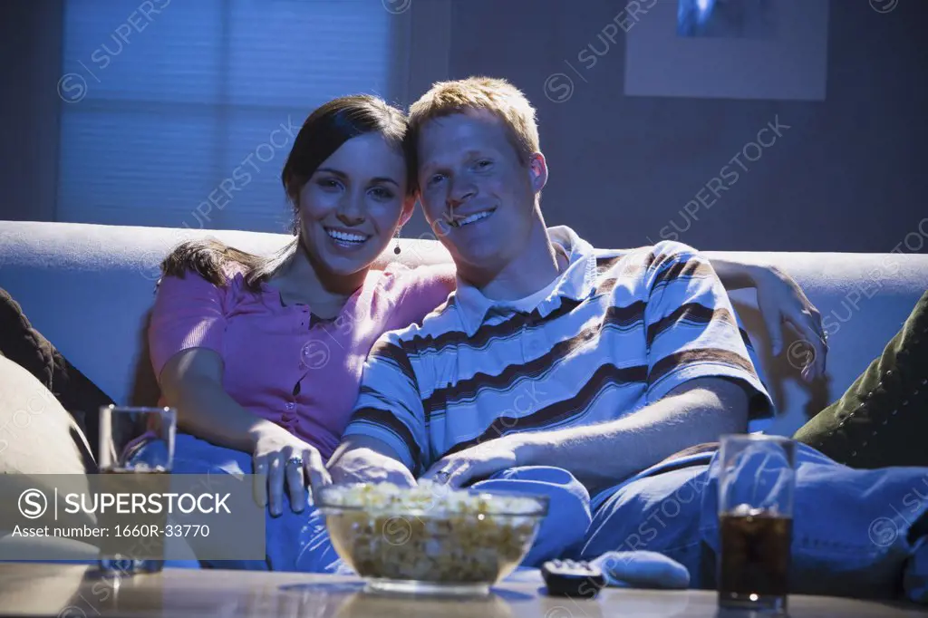 Couple sitting on sofa watching television with bowl of popcorn smiling