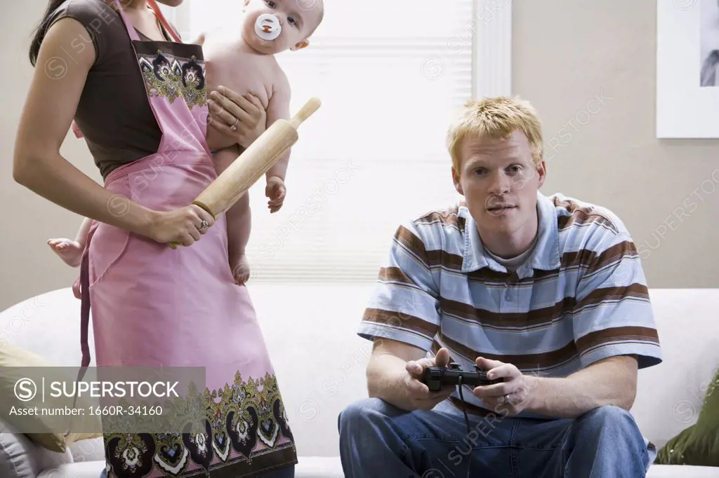 Man on sofa with video game controller and woman in apron with rolling pin holding baby