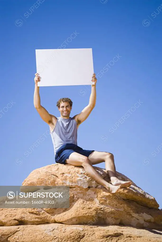 Low angle view of a young man sitting on a rock with a blank sign holding up