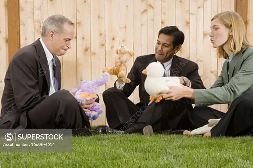 Two businessmen and a businesswoman playing with toys sitting on a lawn