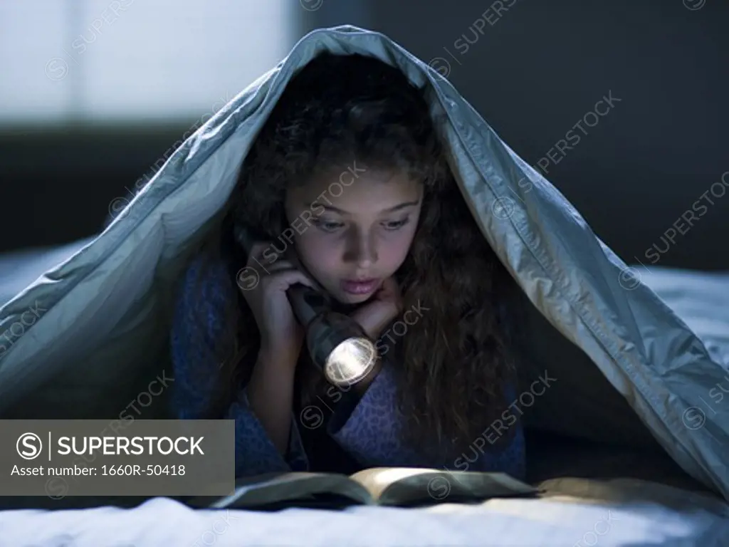 Girl laying in bed under blanket with flashlight reading