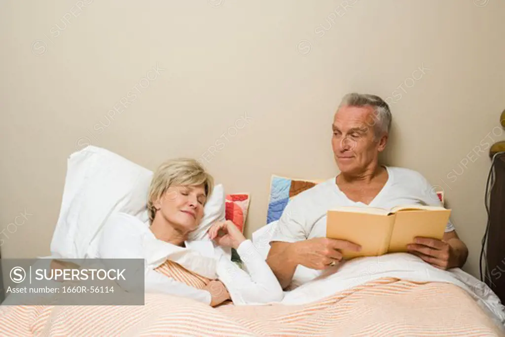 Mature man in bed reading with sleeping woman