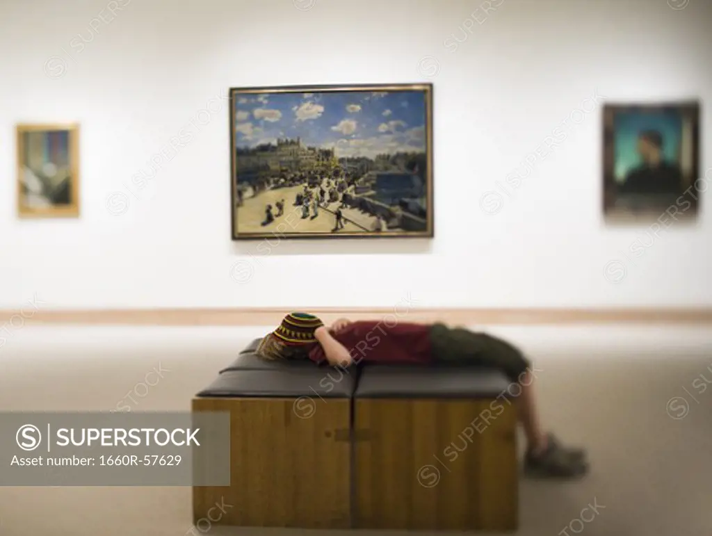 Boy laying down on bench in art gallery with hat covering face