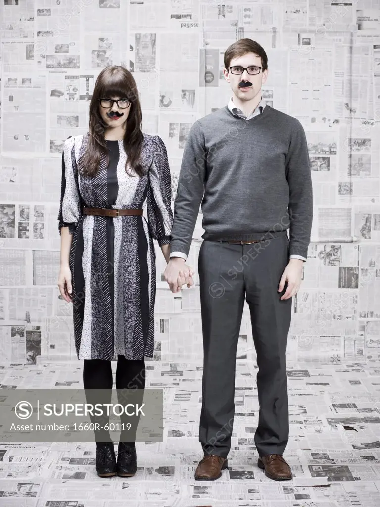 Two young people with fake mustache standing in newspapers covered room, studio shot