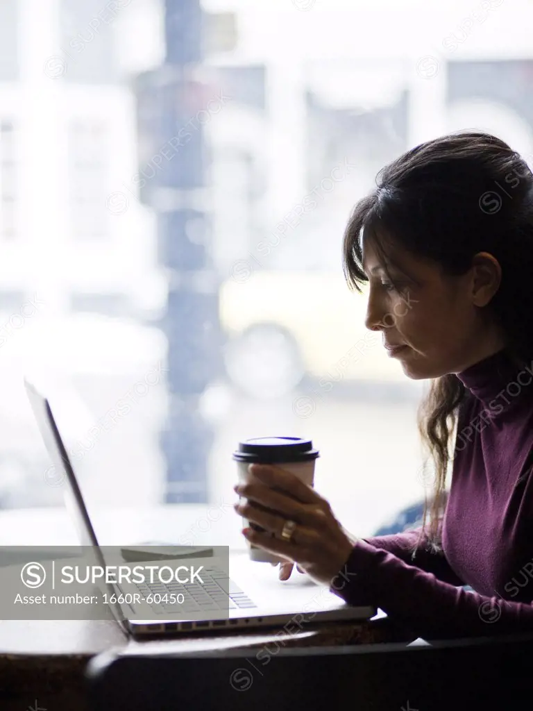 One mid adult woman drinking coffee next to laptop in cafe