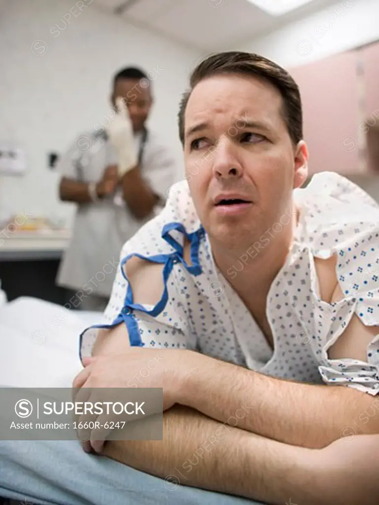 A nervous patient lying on the bed with a doctor putting on latex gloves in the background