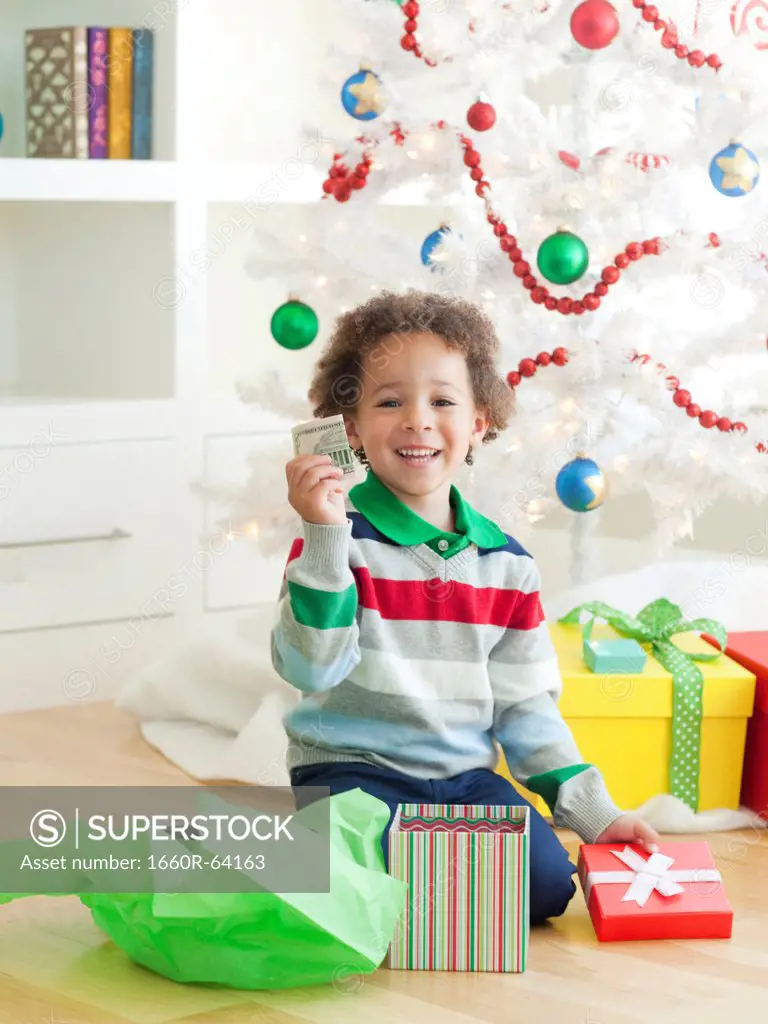 Young boy sitting under Christmas tree, holding dollar note