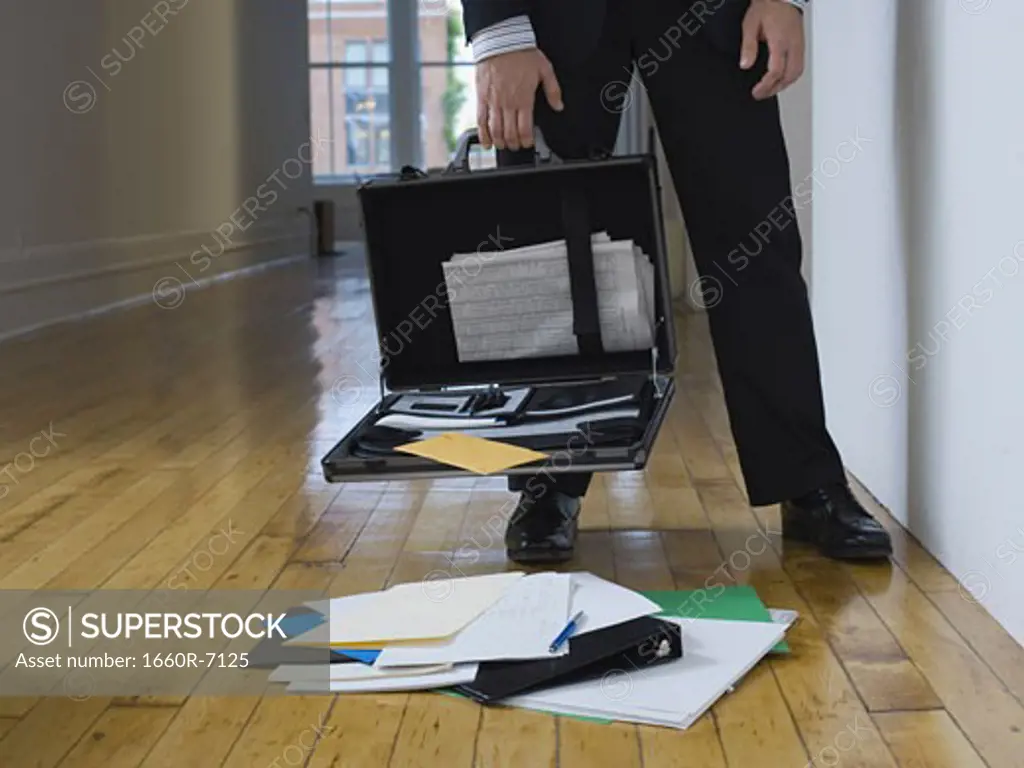 Low section view of a businessman holding an open briefcase with its contents on the floor